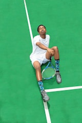 A person wearing a white sports outfit is sitting on a bright green tennis court. The individual has their eyes closed and appears relaxed. They are holding a tennis racket in one hand, and wearing gray sneakers and blue patterned socks.
