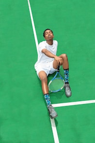 A person wearing a white sports outfit is sitting on a bright green tennis court. The individual has their eyes closed and appears relaxed. They are holding a tennis racket in one hand, and wearing gray sneakers and blue patterned socks.