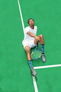 A person wearing a white sports outfit is sitting on a bright green tennis court. The individual has their eyes closed and appears relaxed. They are holding a tennis racket in one hand, and wearing gray sneakers and blue patterned socks.