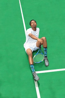 A person wearing a white sports outfit is sitting on a bright green tennis court. The individual has their eyes closed and appears relaxed. They are holding a tennis racket in one hand, and wearing gray sneakers and blue patterned socks.