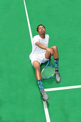 A person wearing a white sports outfit is sitting on a bright green tennis court. The individual has their eyes closed and appears relaxed. They are holding a tennis racket in one hand, and wearing gray sneakers and blue patterned socks.