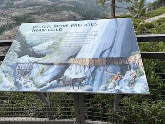 An informational panel detailing the importance of water during the Gold Rush era. It includes an illustration of a wooden aqueduct system being constructed by workers. In the background, a mountainous landscape with trees and rocks is visible.