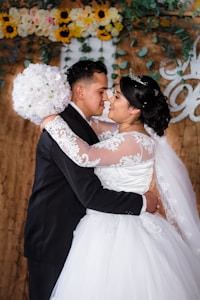 A bride and groom are embracing closely in what appears to be a wedding ceremony. The bride is wearing a white dress with lace details and a tiara, while the groom is dressed in a black suit. The bride is holding a bouquet of white flowers. There is a floral arrangement with yellow and white flowers and green leaves in the background, along with a decorative backdrop featuring some text.