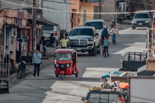 A busy street scene in a small town with various vehicles such as a red auto rickshaw, motorcycles, and cars including a large black pickup truck. Several people are walking and standing on the sidewalks and street. There are buildings with various states of upkeep, and some shops can be seen with items displayed outside.