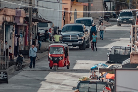 A busy street scene in a small town with various vehicles such as a red auto rickshaw, motorcycles, and cars including a large black pickup truck. Several people are walking and standing on the sidewalks and street. There are buildings with various states of upkeep, and some shops can be seen with items displayed outside.