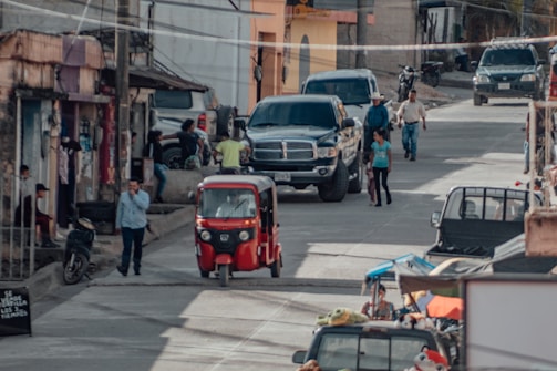 A busy street scene in a small town with various vehicles such as a red auto rickshaw, motorcycles, and cars including a large black pickup truck. Several people are walking and standing on the sidewalks and street. There are buildings with various states of upkeep, and some shops can be seen with items displayed outside.