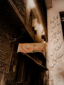 A narrow alleyway with aged brick and plaster walls, partially illuminated by diffuse sunlight coming from an overhead gap. A faded hanging sign with Urdu script is present in the center, along with old windows and tangled electrical wires. The right wall features more Urdu script graffiti-like text.