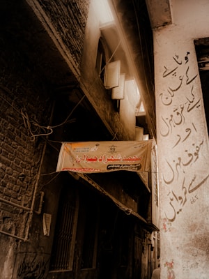 A narrow alleyway with aged brick and plaster walls, partially illuminated by diffuse sunlight coming from an overhead gap. A faded hanging sign with Urdu script is present in the center, along with old windows and tangled electrical wires. The right wall features more Urdu script graffiti-like text.