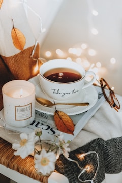 A cozy setting featuring a cup of tea with the phrase 'Take your time' printed on it, placed on a saucer with a golden spoon. Nearby, a lit candle labeled 'TERAZZO' adds warmth and illumination. A single autumn leaf is placed on the saucer, complementing the neutral-toned background. There are folded glasses and a magazine partially visible underneath. A small vase of delicate white flowers adds to the serene ambience, with soft fairy lights in the background enhancing the cozy atmosphere.