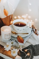 A cozy setting featuring a cup of tea with the phrase 'Take your time' printed on it, placed on a saucer with a golden spoon. Nearby, a lit candle labeled 'TERAZZO' adds warmth and illumination. A single autumn leaf is placed on the saucer, complementing the neutral-toned background. There are folded glasses and a magazine partially visible underneath. A small vase of delicate white flowers adds to the serene ambience, with soft fairy lights in the background enhancing the cozy atmosphere.