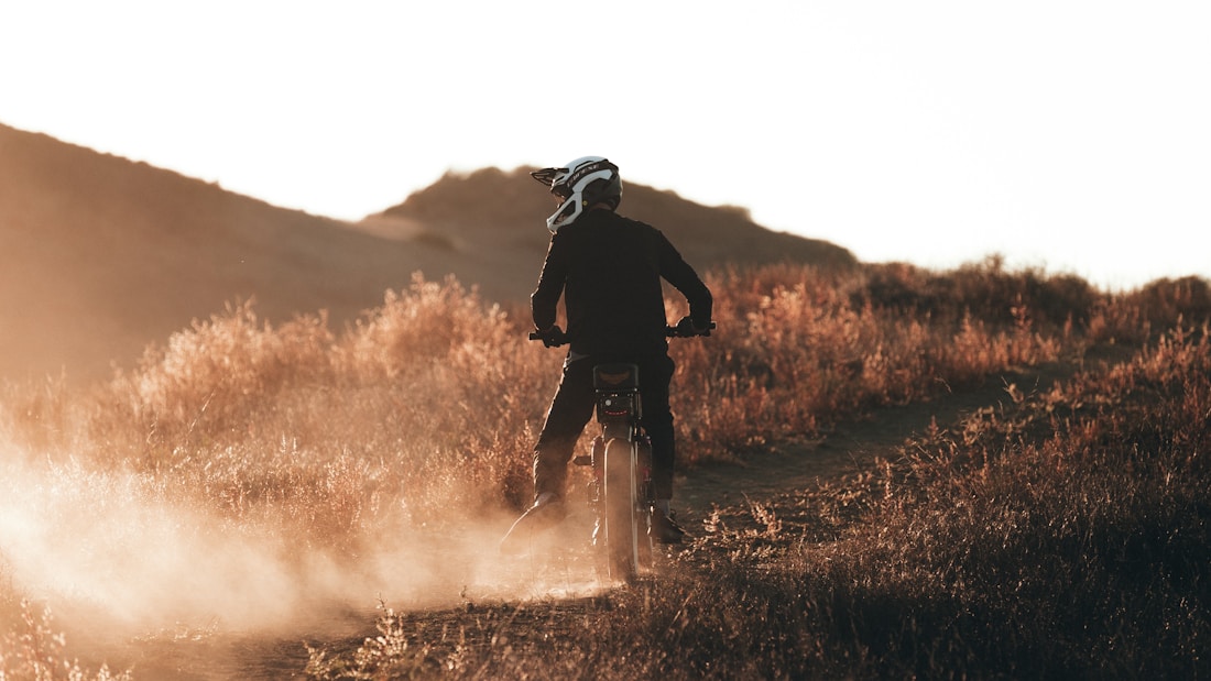 A person rides a dirt bike on a narrow path through a grassy landscape, kicking up dust as they go. The scene is illuminated by a warm, golden light, likely from a setting or rising sun, casting long shadows.
