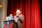 A man is speaking into a microphone at a podium with a decorative emblem, standing on a stage with red curtains in the background. Another person is partially visible behind him, standing near a laptop.