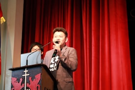 A man is speaking into a microphone at a podium with a decorative emblem, standing on a stage with red curtains in the background. Another person is partially visible behind him, standing near a laptop.