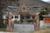 A traditional Bhutanese gate adorned with intricate and colorful designs stands prominently in front of a white building. The gate features vibrant artwork, including dragons and abstract patterns, with a mountainous backdrop. Lush greenery surrounds the area, adding to the serene atmosphere.