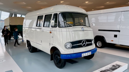 An indoor exhibition featuring a classic white van with a vintage Mercedes emblem. The van is prominently displayed with its front facing the viewer. Next to it, a modern white van is partially visible. The setting appears to be a museum or showroom with polished floors and large windows. Two people, a child and an adult, are seen walking in the background.