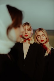 Three women are standing outdoors, each wearing dark jackets and headscarves tied under the chin. The focus is soft, giving a dreamlike, artistic effect. Two women are in sharp focus, with red lipstick complementing their lip color. The foreground features a blurred figure, adding depth to the composition.