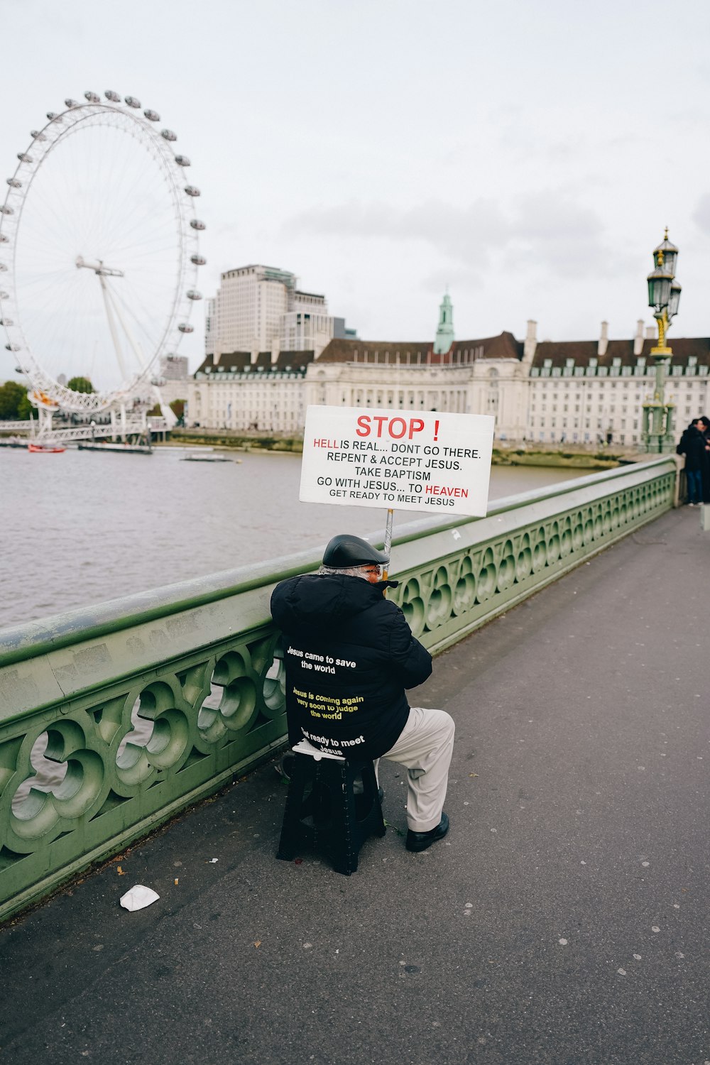 a person kneeling on a bridge with a sign in front of a ferris wheel