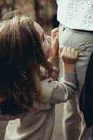 A young child with long brown hair, wearing a gray sweater, is holding on to the pants pocket of an adult. The adult is dressed in light-colored pants and a patterned shirt. The child's facial expression appears to be fond and attentive as they look upwards.