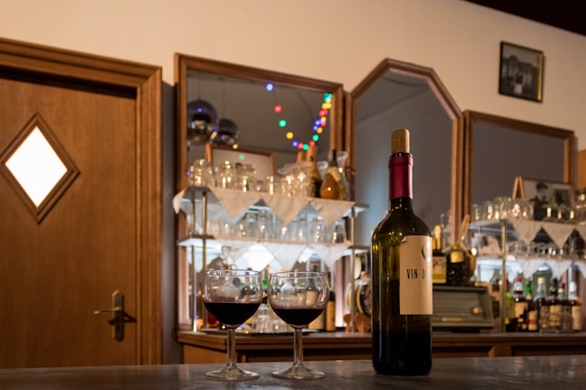 A cozy bar setting featuring a wine bottle and two filled glasses on a wooden counter. Behind them, mirrored shelves display an assortment of glasses, bottles, and small decorative lights.