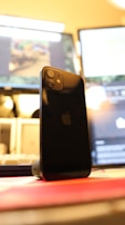 A smartphone is standing upright on a wireless charging stand with a blurred background featuring computer monitors. The phone's back is facing the viewer and prominently displays the manufacturer's logo. The lighting is warm, creating a relaxed atmosphere.