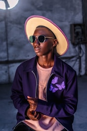 A stylish person wears a large, brimmed hat with a purple trim, glasses, and a dark jacket featuring purple butterfly designs over a light pink shirt. The individual is set against an industrial-style background.