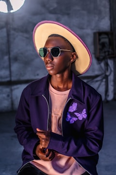 A stylish person wears a large, brimmed hat with a purple trim, glasses, and a dark jacket featuring purple butterfly designs over a light pink shirt. The individual is set against an industrial-style background.