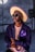 A stylish person wears a large, brimmed hat with a purple trim, glasses, and a dark jacket featuring purple butterfly designs over a light pink shirt. The individual is set against an industrial-style background.