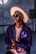 A stylish person wears a large, brimmed hat with a purple trim, glasses, and a dark jacket featuring purple butterfly designs over a light pink shirt. The individual is set against an industrial-style background.