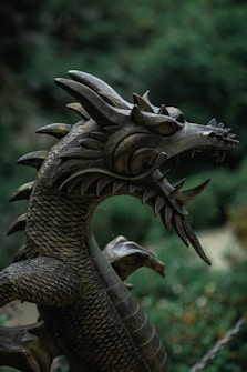 A detailed view of a metallic dragon sculpture, showcasing intricate scale patterns and sharp features. The head is adorned with fierce eyes and distinct spikes, exemplifying a mythical and majestic creature set against a blurred natural background.