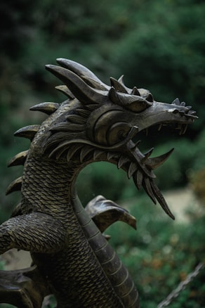 A detailed view of a metallic dragon sculpture, showcasing intricate scale patterns and sharp features. The head is adorned with fierce eyes and distinct spikes, exemplifying a mythical and majestic creature set against a blurred natural background.