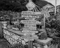 A rustic wooden sign is placed in a garden area with multiple planks displaying messages urging visitors to adhere to specific rules, such as staying on the road, keeping dogs on leads, and enjoying their visit. The background includes stone walls and foliage, contributing to a quaint, countryside atmosphere.