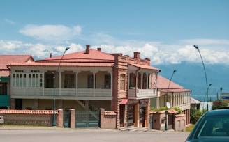 A large two-story brick building with ornate wooden balconies and red roofs is situated on a street. The building features several windows and decorative trim, and a green lawn with some plants is visible in the foreground. Streetlamps line the sidewalk, and the background reveals a mountainous landscape under a partly cloudy sky.