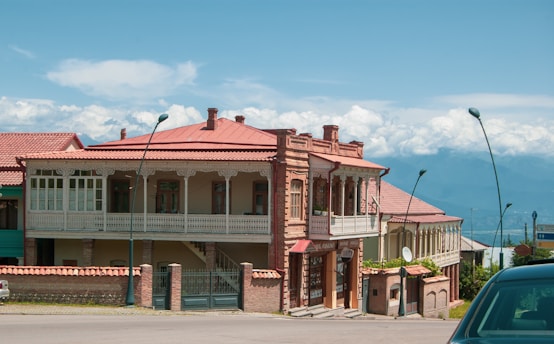 A large two-story brick building with ornate wooden balconies and red roofs is situated on a street. The building features several windows and decorative trim, and a green lawn with some plants is visible in the foreground. Streetlamps line the sidewalk, and the background reveals a mountainous landscape under a partly cloudy sky.
