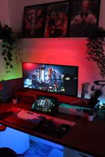 A modern gaming setup is illuminated by ambient lighting. A large monitor displays a futuristic cityscape, accompanied by a tablet and smartphone showing the same image. A mechanical keyboard and a microphone are on the desk, surrounded by small potted plants and gaming accessories. Vibrant red and blue lights create a cyberpunk atmosphere, with posters of anime characters hanging above.