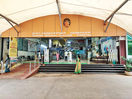 Entrance of a medical college and hospital with a large archway displaying the name Tagore Medical College & Hospital. People are walking towards the entrance, with some sitting on a bench and others interacting. Signage and informational posters are visible on the walls, and wheelchairs are parked nearby. There is a ramp and steps leading up to the entrance.