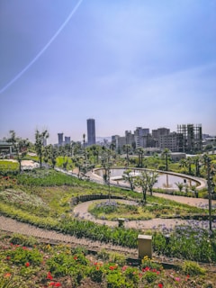 A landscaped park with winding paths and vibrant flowerbeds situated in an urban environment. There are tall buildings in the background, and a prominent curved water feature surrounded by greenery. Trees are scattered throughout the park area.