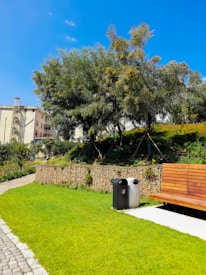 A well-maintained park area featuring a wooden bench on a concrete slab surrounded by lush green grass. Tall trees with dense foliage provide shade, and a retaining wall made of stacked stones runs along the edge. Two modern trash bins, one black and one silver, are situated near the bench. A building with multiple windows can be seen in the background under a clear blue sky.