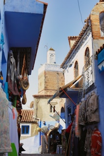A narrow alley lined with colorful textiles and traditional goods on display, leading towards a stone building with a tower. The architecture is characterized by blue and white painted structures with intricate designs, creating a charming and vibrant scene. People are walking through the alley, adding life to the market atmosphere.