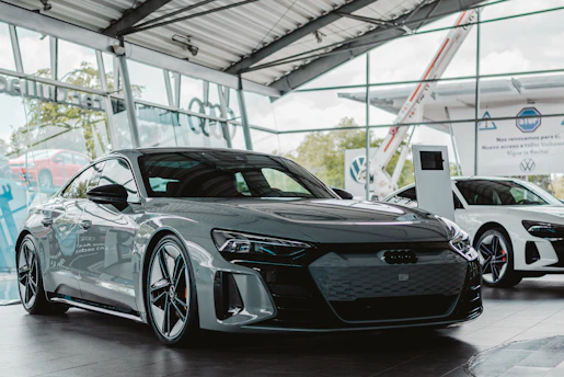 A sleek, modern car is displayed in a showroom with large glass windows. The car is a glossy dark color, featuring a low profile and sharp, aerodynamic lines. In the background, another white car is visible alongside digital display screens and signage, giving the space a futuristic and luxurious atmosphere.