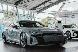 A sleek, modern car is displayed in a showroom with large glass windows. The car is a glossy dark color, featuring a low profile and sharp, aerodynamic lines. In the background, another white car is visible alongside digital display screens and signage, giving the space a futuristic and luxurious atmosphere.