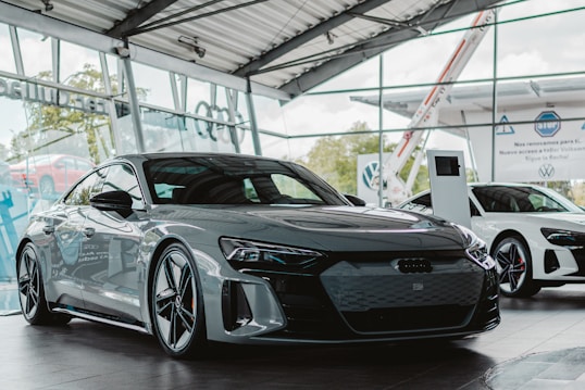 A sleek, modern car is displayed in a showroom with large glass windows. The car is a glossy dark color, featuring a low profile and sharp, aerodynamic lines. In the background, another white car is visible alongside digital display screens and signage, giving the space a futuristic and luxurious atmosphere.