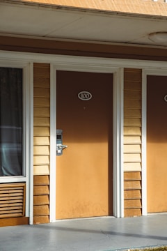 The image shows a section of a building exterior with a brown door numbered 100. The door is flanked by vertical wooden siding in light beige and dark brown colors. To the left of the door, a window is partially covered by a curtain, and below the window is a vent or air conditioning unit. The ground outside the door is paved, and the ceiling above has a light fixture.