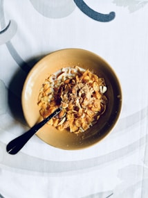 A ceramic bowl filled with cornflakes and milk is placed at the center of a patterned tablecloth. Almonds and a sprinkle of powdered sugar are added on top of the cereal. A spoon is placed inside the bowl, suggesting it is ready to be eaten.