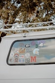 The back window of a white vehicle is decorated with various stickers, each representing different universities, teams, or organizations. The surrounding area includes tree branches and leaves.