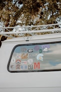 The back window of a white vehicle is decorated with various stickers, each representing different universities, teams, or organizations. The surrounding area includes tree branches and leaves.