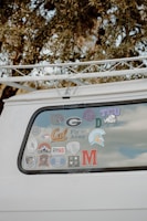 The back window of a white vehicle is decorated with various stickers, each representing different universities, teams, or organizations. The surrounding area includes tree branches and leaves.