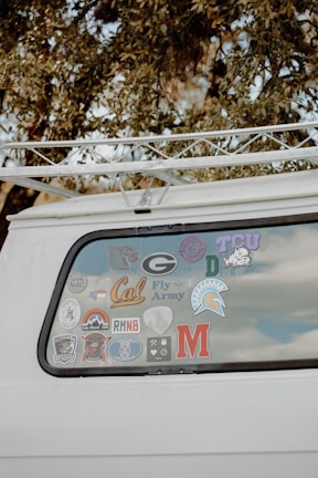 The back window of a white vehicle is decorated with various stickers, each representing different universities, teams, or organizations. The surrounding area includes tree branches and leaves.