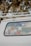 The back window of a white vehicle is decorated with various stickers, each representing different universities, teams, or organizations. The surrounding area includes tree branches and leaves.