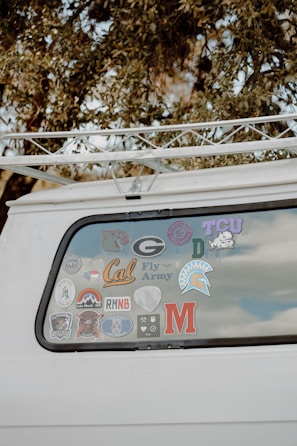 The back window of a white vehicle is decorated with various stickers, each representing different universities, teams, or organizations. The surrounding area includes tree branches and leaves.