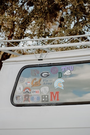 The back window of a white vehicle is decorated with various stickers, each representing different universities, teams, or organizations. The surrounding area includes tree branches and leaves.